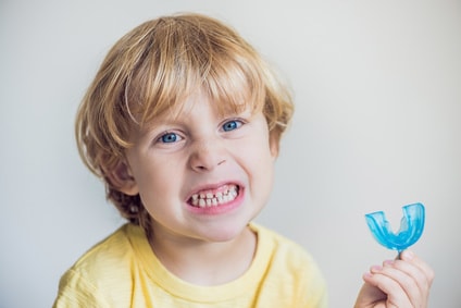 Ein kleines Kind zeigt seine Zähne und hält eine Zahnspangenschiene in der Hand. Schon bei kleinen Kindern kann eine kieferorthopädische Behandlung notwendig sein.